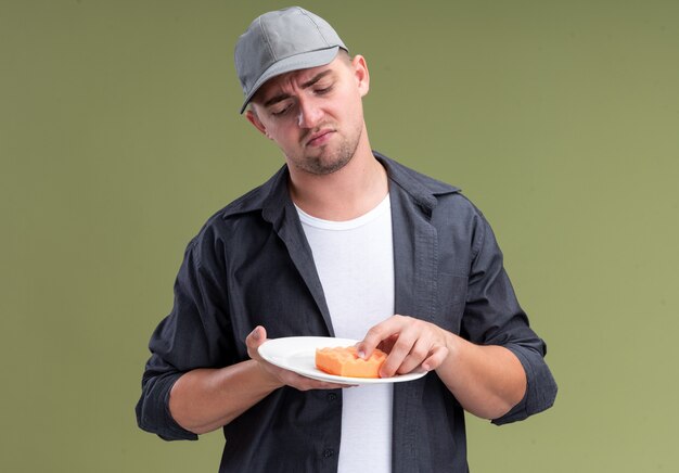 Недовольный молодой красивый уборщик в футболке и кепке, держащий тарелку с губкой, изолированную на оливково-зеленой стене