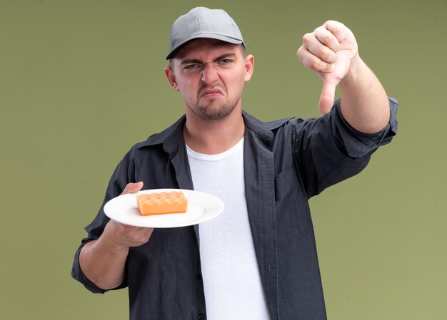 Unpleased young handsome cleaning guy wearing t-shirt and cap holding sponge on plate showing thumb down isolated on olive green wall