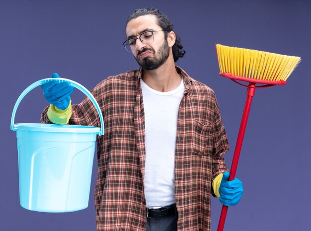 Unpleased young handsome cleaning guy wearing t-shirt and gloves holding bucket with mop isolated on blue wall