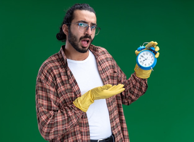 Unpleased young handsome cleaning guy wearing t-shirt and gloves holding and points at alarm clock isolated on green wall