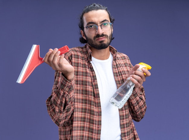 Unpleased young handsome cleaning guy wearing t-shirt holding mop head with spray bottle isolated on blue wall