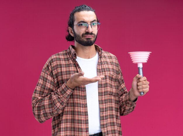 Unpleased young handsome cleaning guy wearing t-shirt holding and points at plunger isolated on pink wall