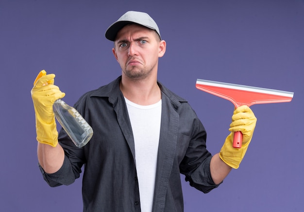 Unpleased young handsome cleaning guy wearing t-shirt with cap and gloves holding mop head and spray bottle isolated on purple wall