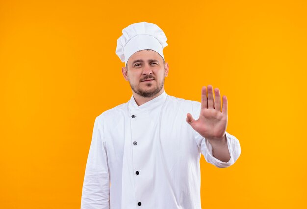 Unpleased young handsome cook in chef uniform gesturing stop isolated on orange space