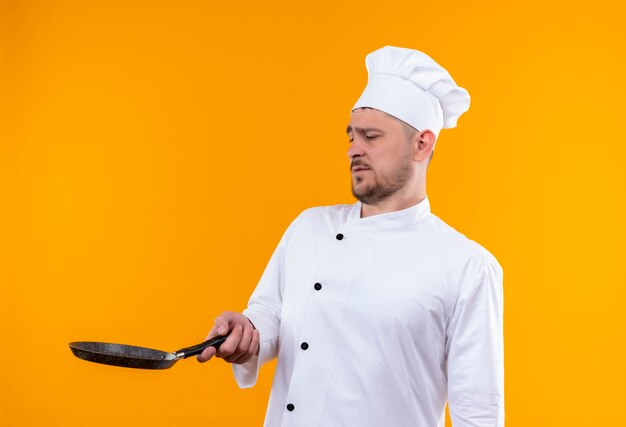 Unpleased young handsome cook in chef uniform holding and looking at frying pan isolated on orange space