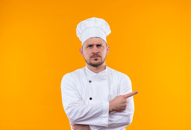 Недовольный молодой красивый повар в униформе шеф-повара, указывая в сторону на изолированном оранжевом пространстве