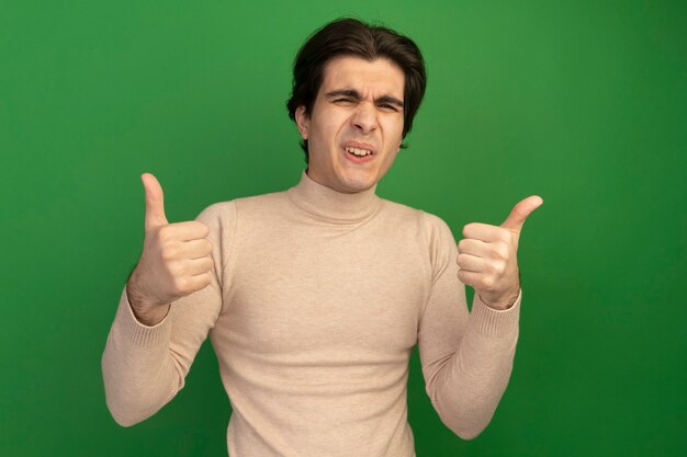 Unpleased young handsome guy showing thumbs up isolated on green wall