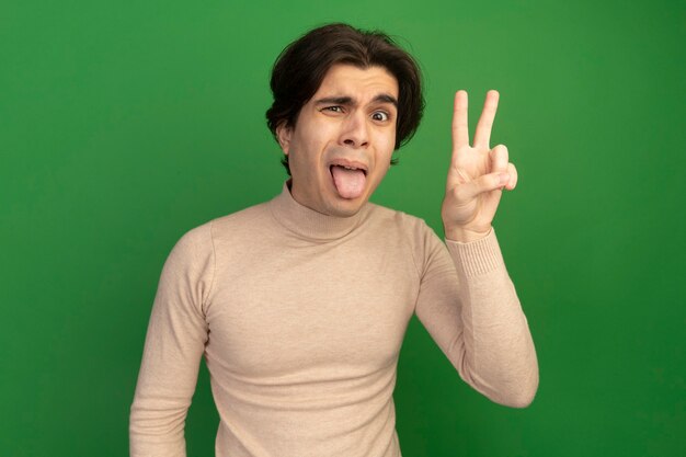 Бесплатное фото Недовольный молодой красивый парень показывает язык, делая жест мира, изолированный на зеленой стене
