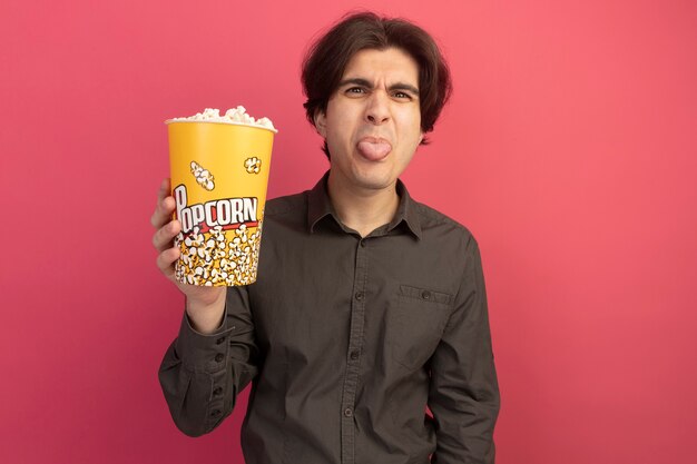 Unpleased young handsome guy wearing black t-shirt holding bucket of popcorn and showing tongue isolated on pink wall