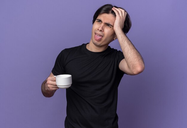Недовольный молодой красивый парень в черной футболке держит чашку кофе, показывает язык и кладет руку на голову, изолированную на фиолетовой стене