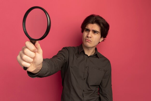 Unpleased young handsome guy wearing black t-shirt holding and looking at magnifier isolated on pink wall