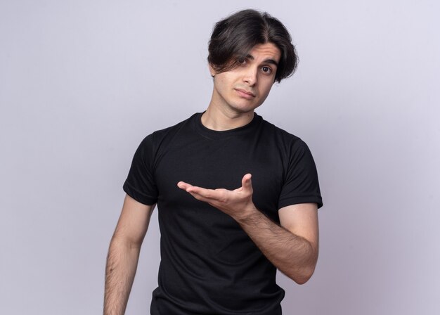 Unpleased young handsome guy wearing black t-shirt points with hand at side isolated on white wall