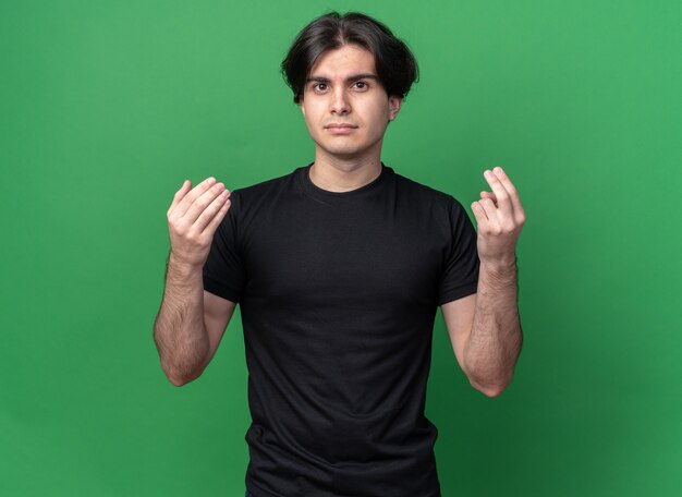 Недовольный молодой красивый парень в черной футболке, показывающий жест чаевых, изолированный на зеленой стене