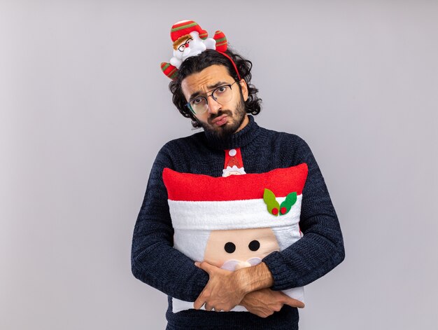 Unpleased young handsome guy wearing christmas tie with hair hoop hugged pillow isolated on white background