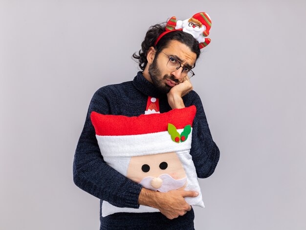 Unpleased young handsome guy wearing christmas tie with hair hoop hugged pillow putting hand on cheek isolated on white background