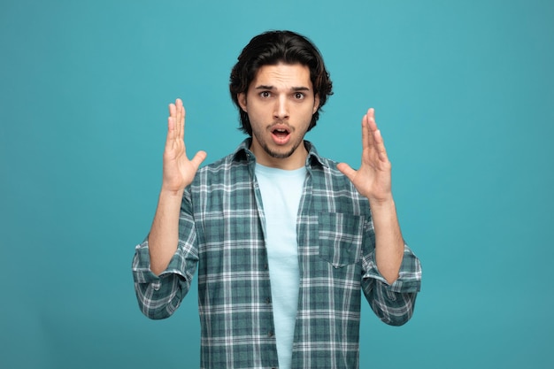 unpleased young handsome man looking at camera keeping hands in air isolated on blue background