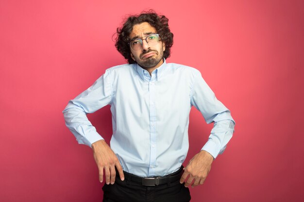 Unpleased young handsome man wearing glasses looking at front keeping hands in air isolated on pink wall