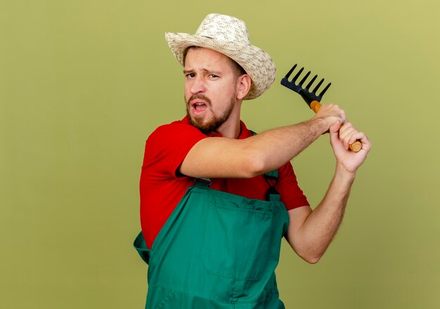Unpleased young handsome slavic gardener in uniform and hat holding rake  getting ready to hit someone with rake isolated on olive green wall with copy space