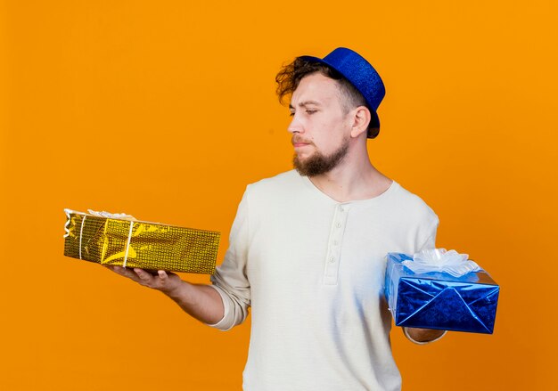 Unpleased young handsome slavic party guy wearing party hat holding and looking at gift boxes isolated on orange background