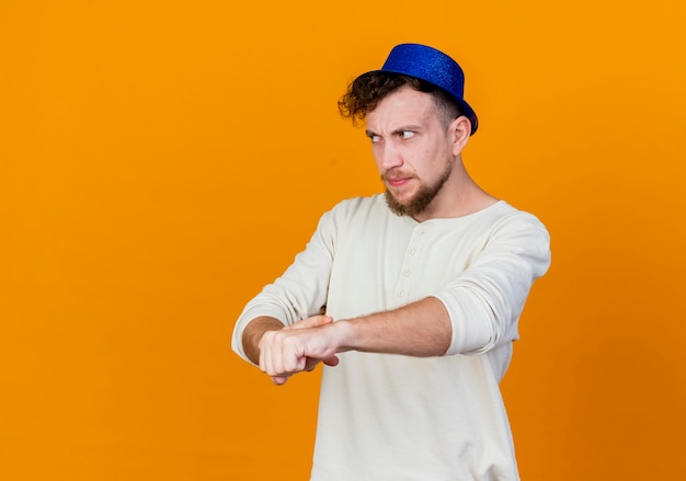 Unpleased young handsome slavic party guy wearing party hat looking straight doing you are late gesture isolated on orange background with copy space