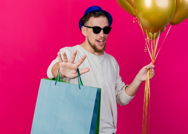 Unpleased young handsome slavic party guy wearing party hat and sunglasses holding balloons and paper bags looking at camera doing no gesture isolated on crimson background