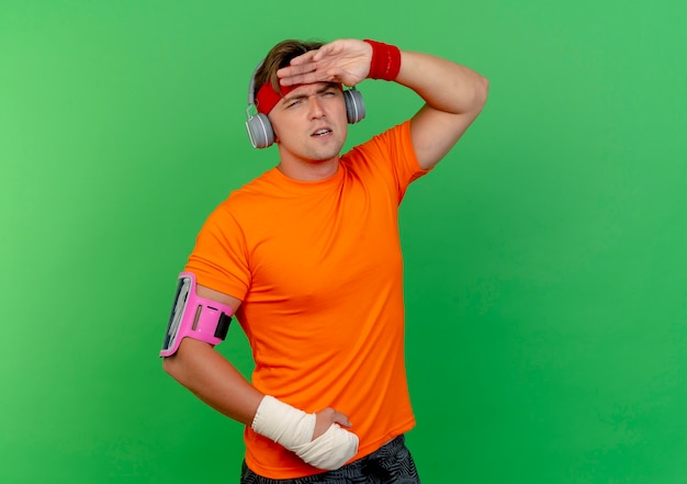 Free Photo unpleased young handsome sporty man wearing headband and wristbands and headphones and phone armband with wrist wrapped with bandage putting hand on forehead looking straight