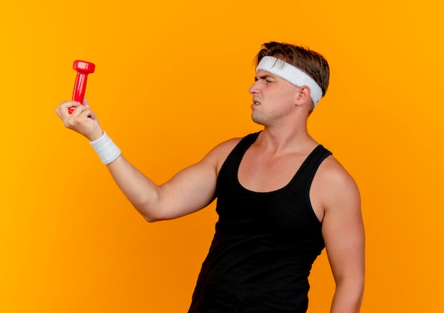 Unpleased young handsome sporty man wearing headband and wristbands holding and looking at dumbbell isolated on orange background