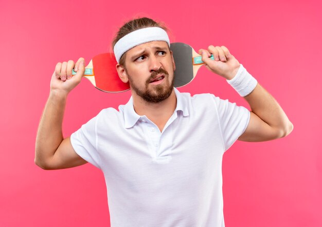 Unpleased young handsome sporty man wearing headband and wristbands holding ping pong rackets behind his head and looking at side isolated on pink space