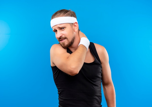 Unpleased young handsome sporty man wearing headband and wristbands putting hand on his neck isolated on blue space 