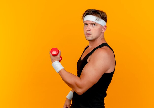 Unpleased young handsome sporty man wearing headband and wristbands standing in profile view holding dumbbell isolated on orange background with copy space