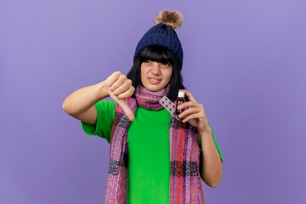 Unpleased young ill caucasian girl wearing winter hat and scarf holding medicament in glass and pack of capsules looking at side showing thumb down isolated on purple background with copy space