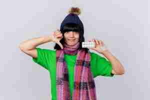 Free photo unpleased young ill caucasian girl wearing winter hat and scarf looking at camera showing pack of tablets and thumb down isolated on white background with copy space