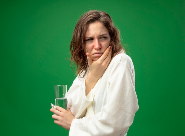 Foto gratuita giovane ragazza malata dispiaciuta che guarda il lato che indossa una veste bianca che tiene il bicchiere d'acqua mettendo la mano sulla guancia isolata sul verde