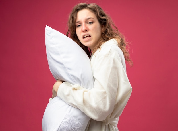 ピンクで隔離の白いローブ抱き枕を身に着けている側を見て不機嫌な若い病気の女の子