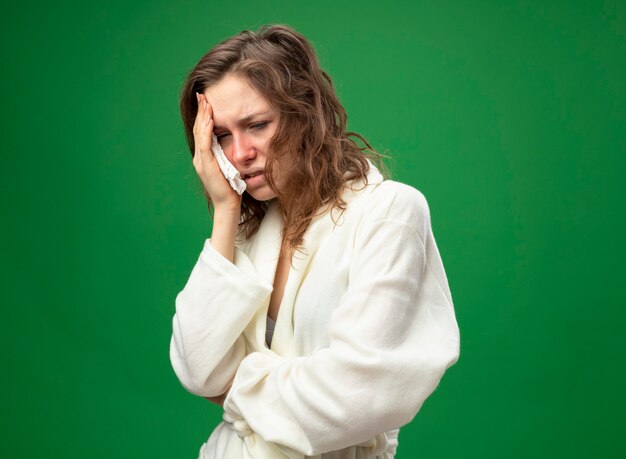 Unpleased young ill girl looking at side wearing white robe putting hand on face isolated on green