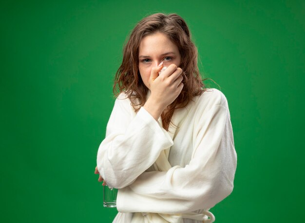 Unpleased young ill girl looking straight ahead wearing white robe putting hand on mouth isolated on green