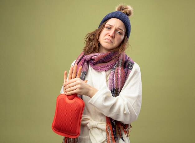 Unpleased young ill girl looking straight ahead wearing white robe and winter hat with scarf holding hot water bag isolated on olive green