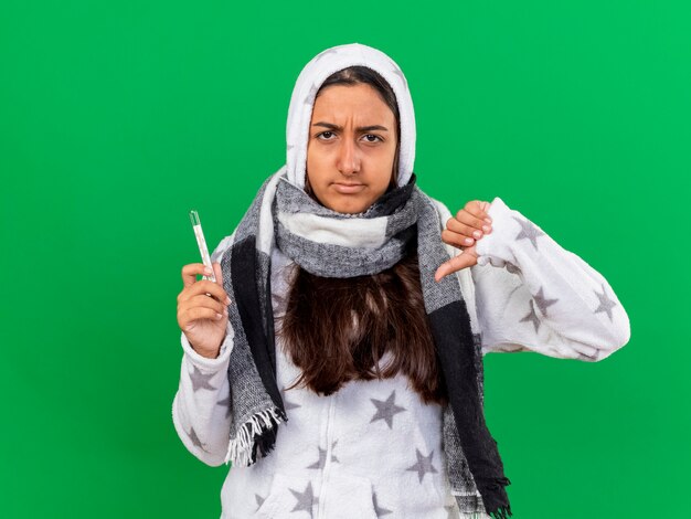 Unpleased young ill girl putting on hood wearin scarf holding thermometer showing thumb down isolated on green