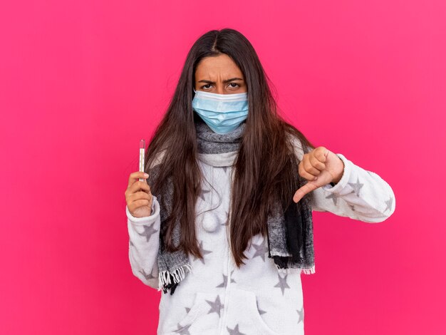 ピンクの背景で隔離の親指を下に示す温度計を保持しているスカーフと医療マスクを身に着けている不機嫌な若い病気の女の子