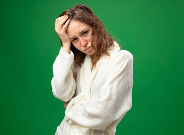 Unpleased young ill girl wearing white robe putting hand on head
