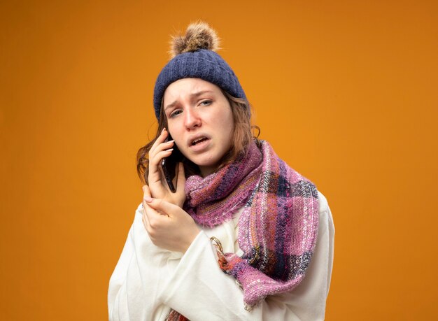 Unpleased young ill girl wearing white robe and winter hat with scarf speaks on phone isolated on orange wall