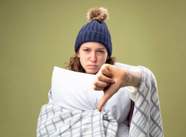 Недовольная молодая больная девушка в белом халате и зимней шапке с шарфом, обернутым в клетчатую подушку, с большим пальцем вниз