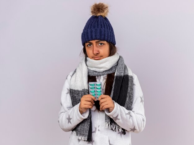 Unpleased young ill girl wearing winter hat with scarf holding pills isolated on white