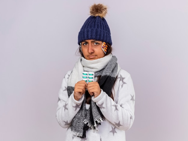 Unpleased young ill girl wearing winter hat with scarf holding pills isolated on white