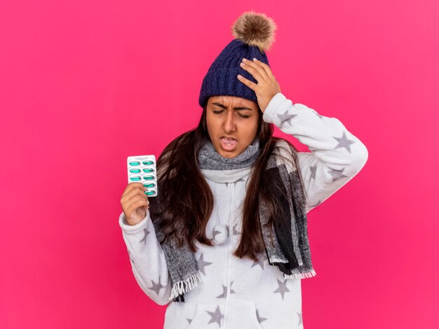 Unpleased young ill girl wearing winter hat with scarf holding pills and putting hand on aching head isolated on pink