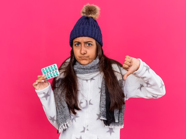 Недовольная молодая больная девушка в зимней шапке с шарфом держит таблетки и показывает большой палец вниз на розовом фоне