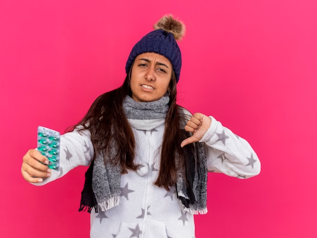 Unpleased young ill girl wearing winter hat with scarf holding pills and showing thumb down isolated on pink