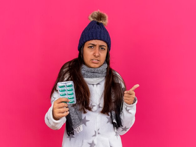 Недовольная молодая больная девушка в зимней шапке с шарфом держит таблетки, показывая вам жест, изолированный на розовом