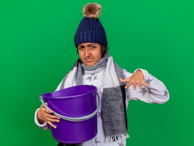 Unpleased young ill girl wearing winter hat with scarf holding plastic bucket isolated on green