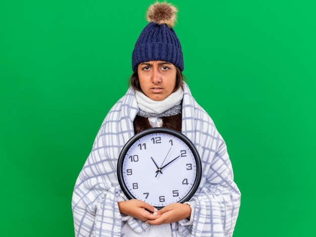 Бесплатное фото Недовольная молодая больная девушка в зимней шапке с шарфом держит настенные часы, изолированные на зеленом фоне